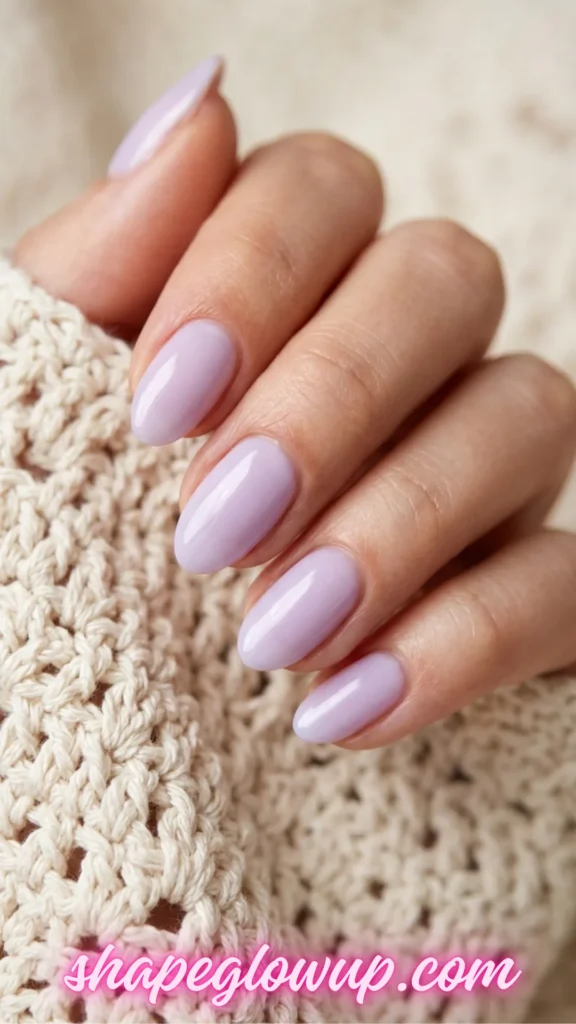 A close-up view of a woman’s hand showcasing a glossy pastel lavender manicure on medium-length, almond-shaped nails. The hand gently rests against a textured, cream-colored knit crochet sleeve to highlight the color pairing.
