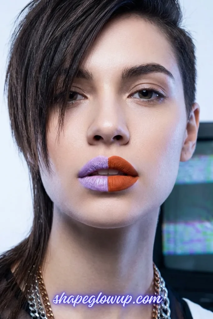 A close-up portrait of a person with a short, edgy dark hairstyle showcasing an intentional warm and cool toned mismatched makeup look. Their lips are split vertically down the center: the right side is painted with a frosted, metallic lavender purple, while the left side features a matte, warm terracotta orange. Layered silver and gold chain necklaces are visible, and a blurred computer monitor with a pixelated glitch graphic sits in the background. A text watermark at the bottom reads "shapeglowup.com".